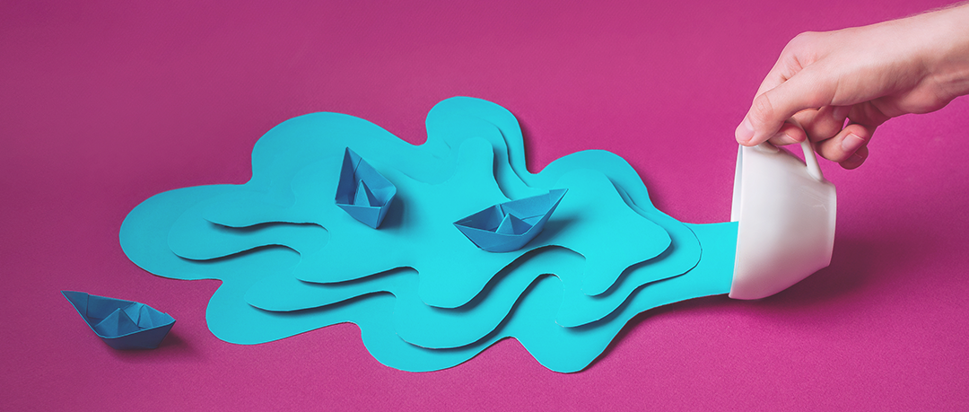 Papercraft Stockfoto: eine Hand kippt eine Tasse aus, heraus fließen Wasser und Schiffchen - beides aus blauem Papier geschnitten und modelliert